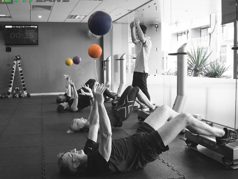 Group exercise class at FitLane. Gym goers lying on floor throwing medicine ball in the air