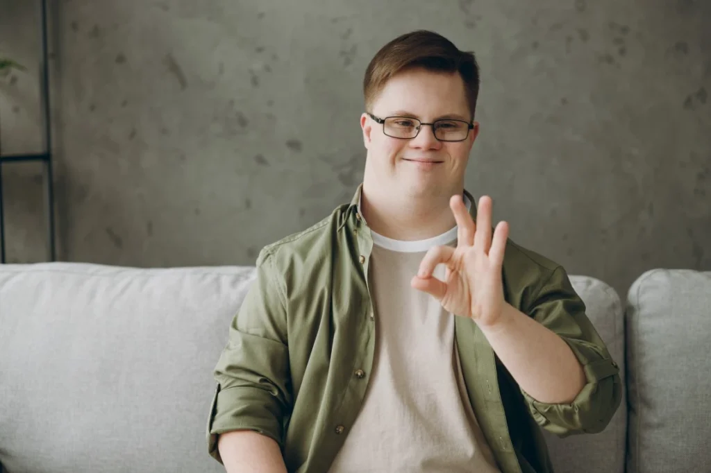 Disabled man with down syndrome sitting on couch giving ok symbol to camera and smiling