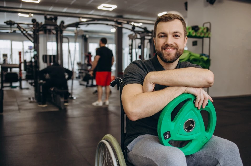 Happy man in wheelchair at the gym holding 10kg weight in lap and smiling at camera