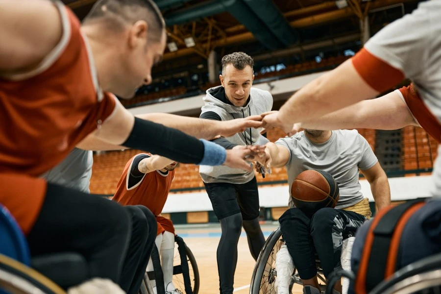 Group of disabled friends in wheelchairs playing basketball, putting hands in centre of group with coach