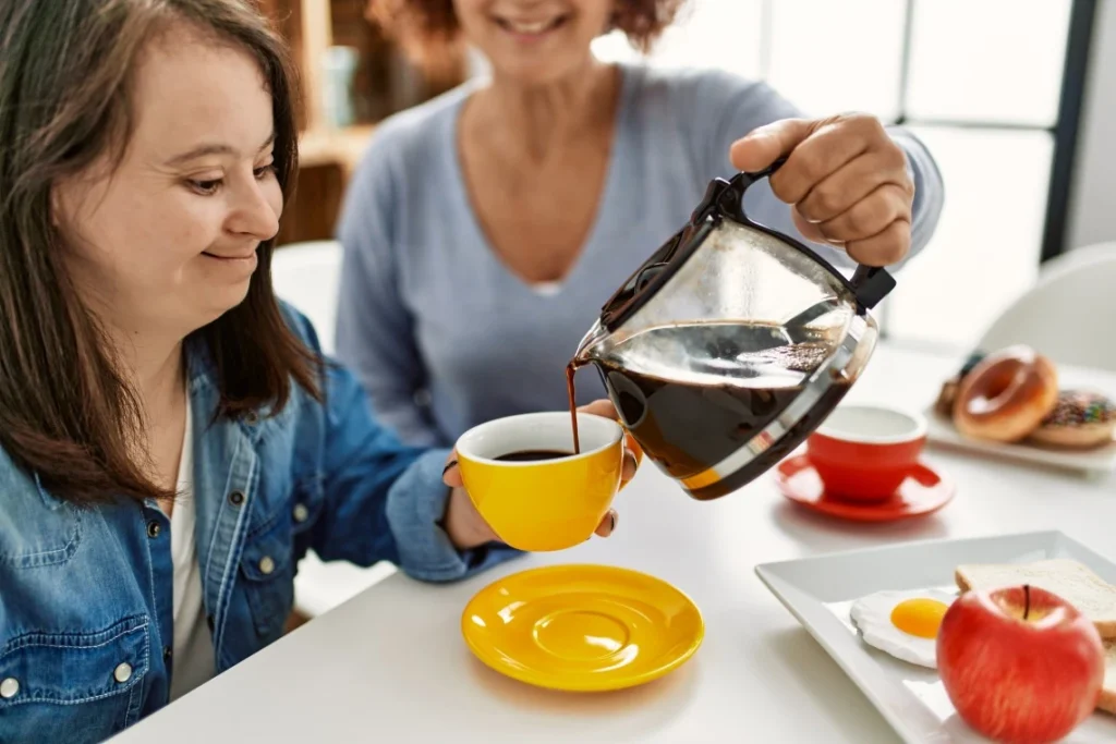 Close up of disabled girl with down syndrome holding coffee mug while support worker pours coffee
