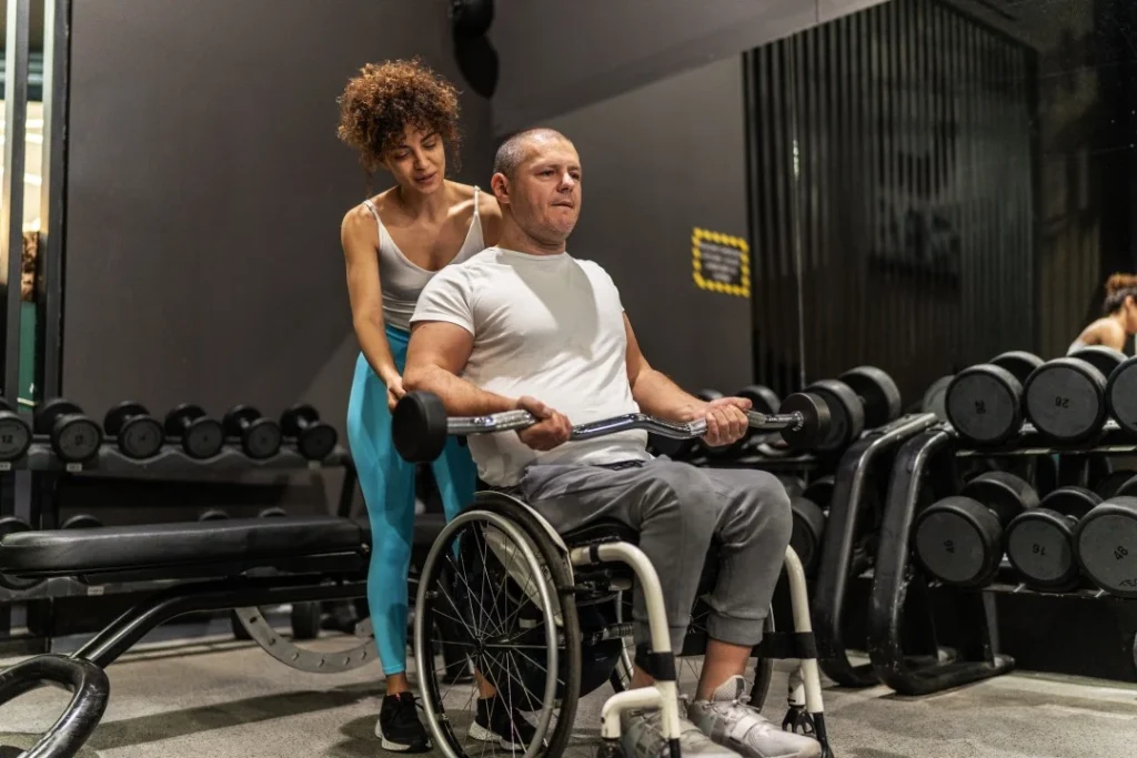 Disabled man in wheelchair in the gym with trainer standing behind him helping to life weight