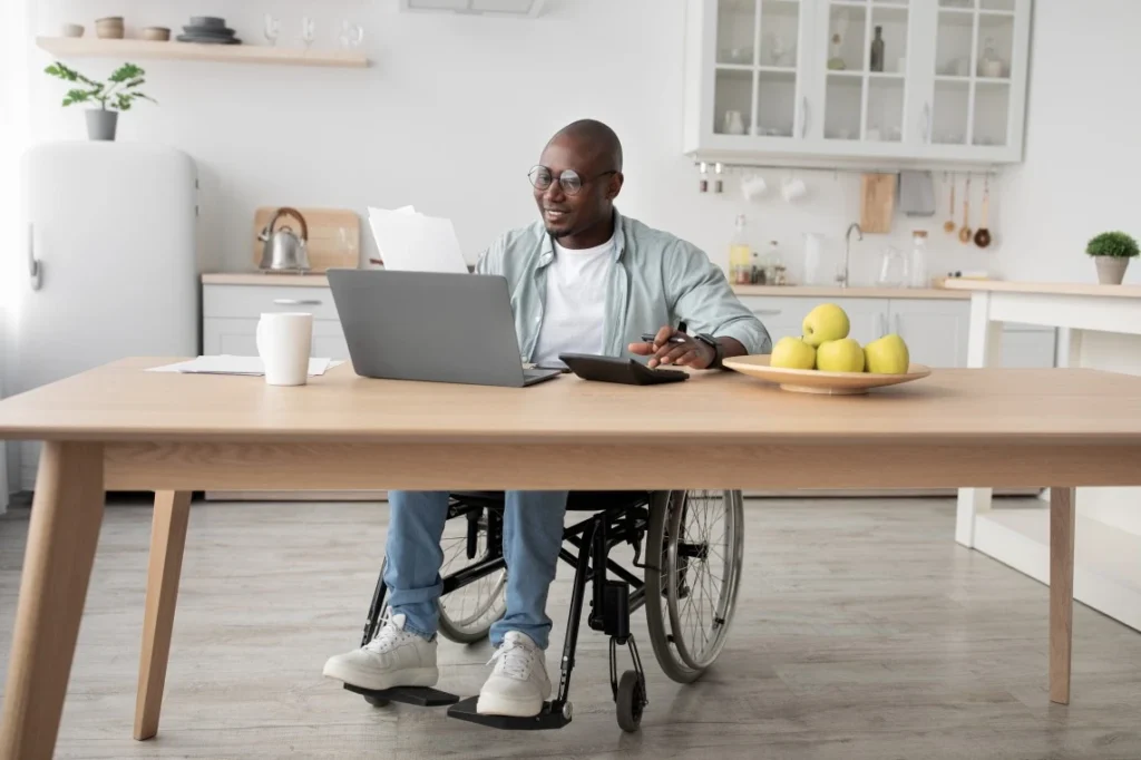 Disabled man in wheelchair sitting at desk looking at laptop and using calculator to budget