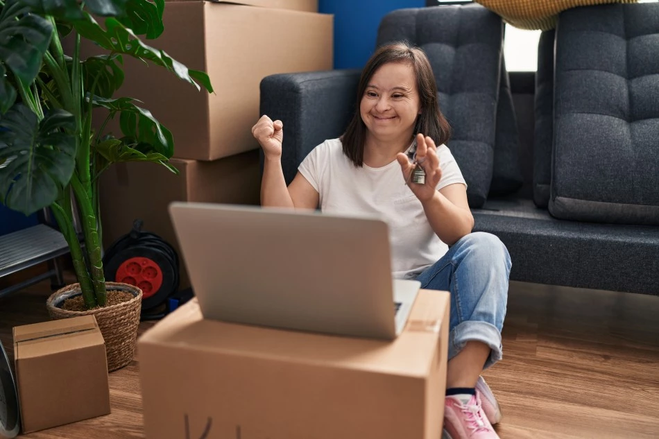 Disabled girl with down syndrome sitting on floor with moving boxes in front of laptop holding keys to new home and celebrating